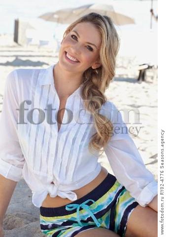 A young blonde woman sitting on a beach wearing a white blouse and colourful shorts