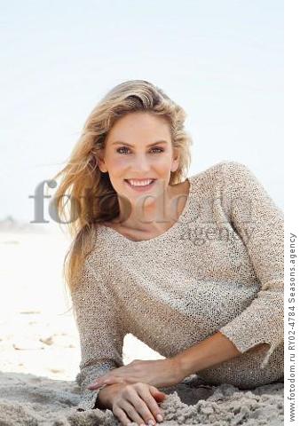 A young blonde woman lying on a beach wearing a beige knitted jumper