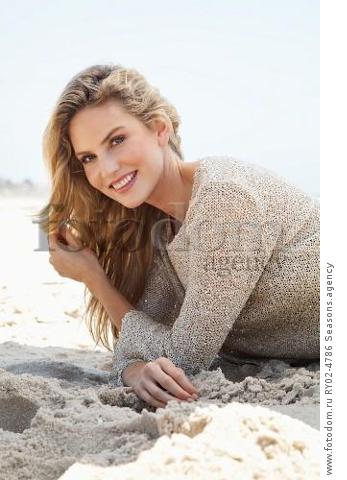 A young blonde woman lying on a beach wearing a beige knitted jumper