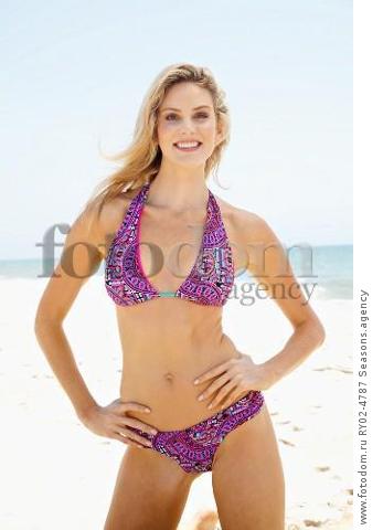 A young blonde woman on a beach wearing a purple bikini with her hands on her hips