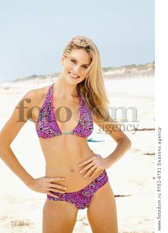 A young blonde woman on a beach wearing a purple bikni with her hands on her hips