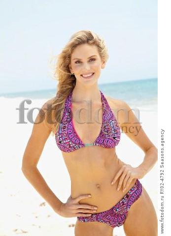A young blonde woman on a beach with her hands on her hips wearing a purple bikini