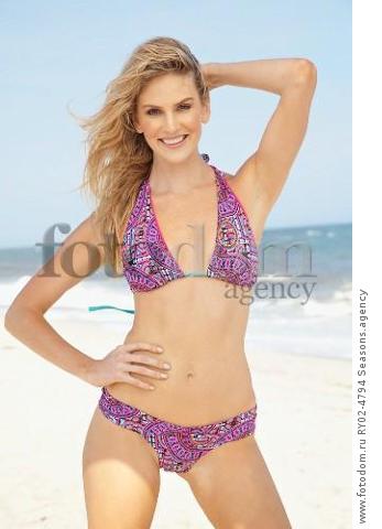 A young blonde woman on a beach with one hand on her hip wearing a purple bikini