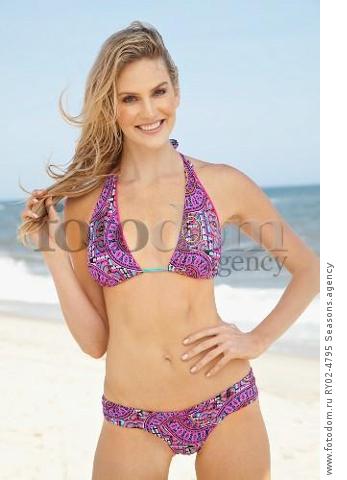 A young blonde woman on a beach wearing a purple bikini with one hand on her hip
