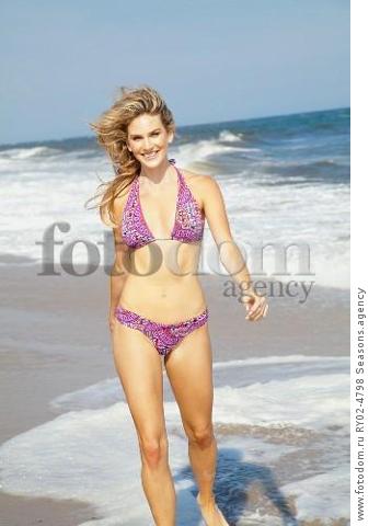 A young blonde woman running on a beach wearing a purple bikini